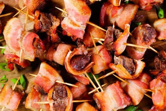 Bacon wrapped steak bites piled on a cutting board with chopped green onion garnish.