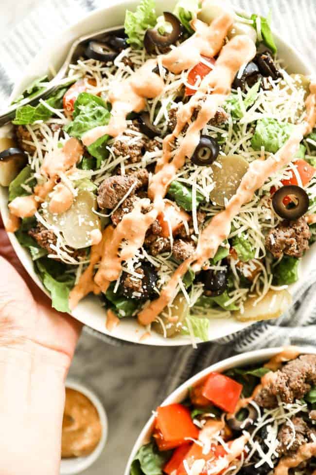 Holding a bowl full of cheeseburger salad with shredded cheese on top and special sauce drizzled all over the top.