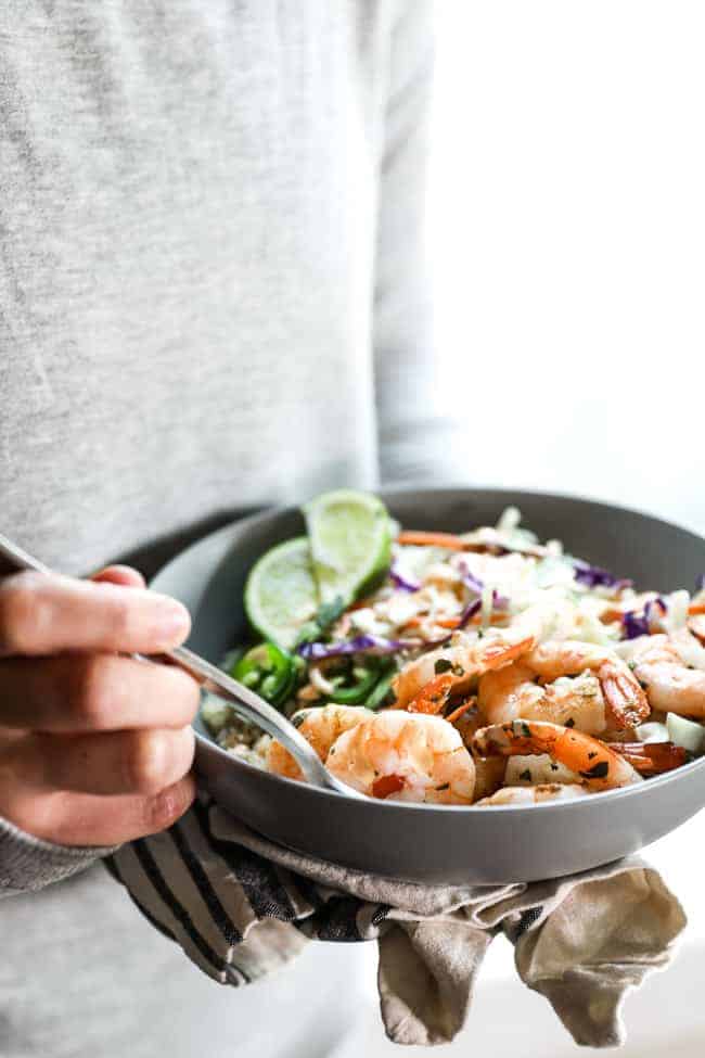 Holding bowl and digging fork into cilantro lime shrimp bowl. Bowl is topped with lime wedges, jalapeño slices and sauce.