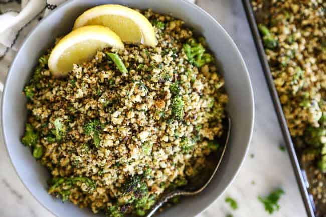 Crispy roasted cauliflower rice in a serving bowl with a spoon and lemon wedges.