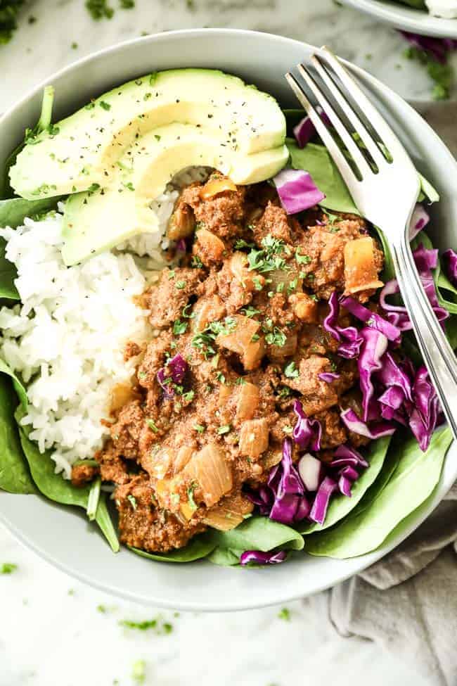 Close up vertical image of instant pot sloppy joe's served in bowl with spinach, white rice, purple cabbage, parsley and avocado. Fork dug into bowl.