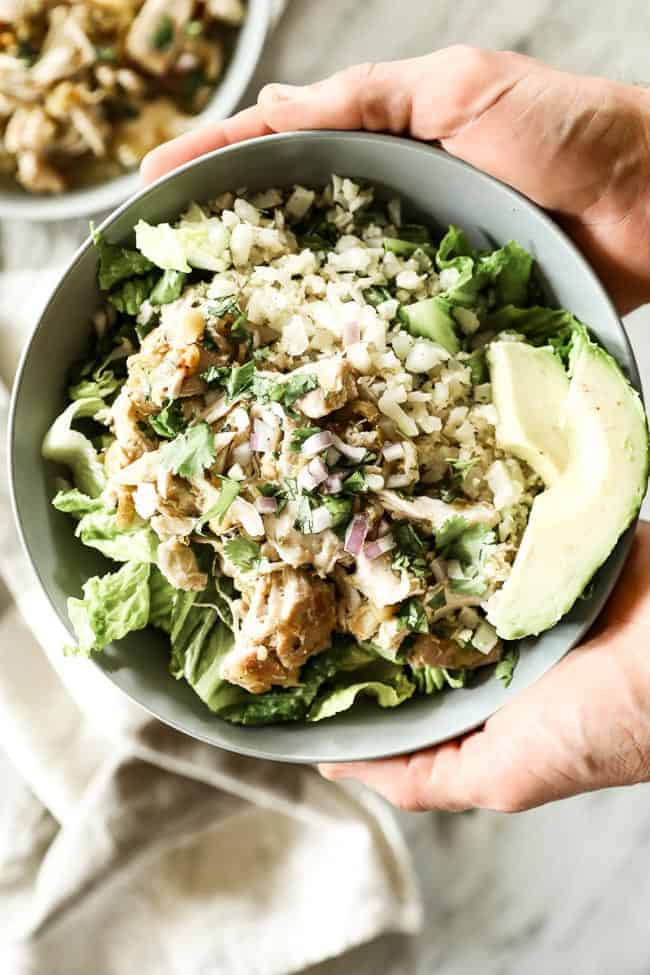 Holding a bowl of Mexican shredded chicken with romaine lettuce, caulirice, shredded chicken, avocado and fresh chopped red onion and cilantro.