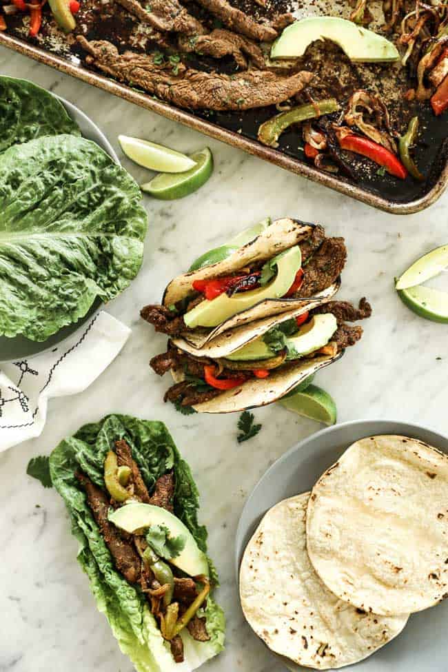 Vertical overhead image of sheet pan fajitas served in soft taco shells and also in romaine lettuce leaves. Topped with sliced avocado and fresh cilantro.