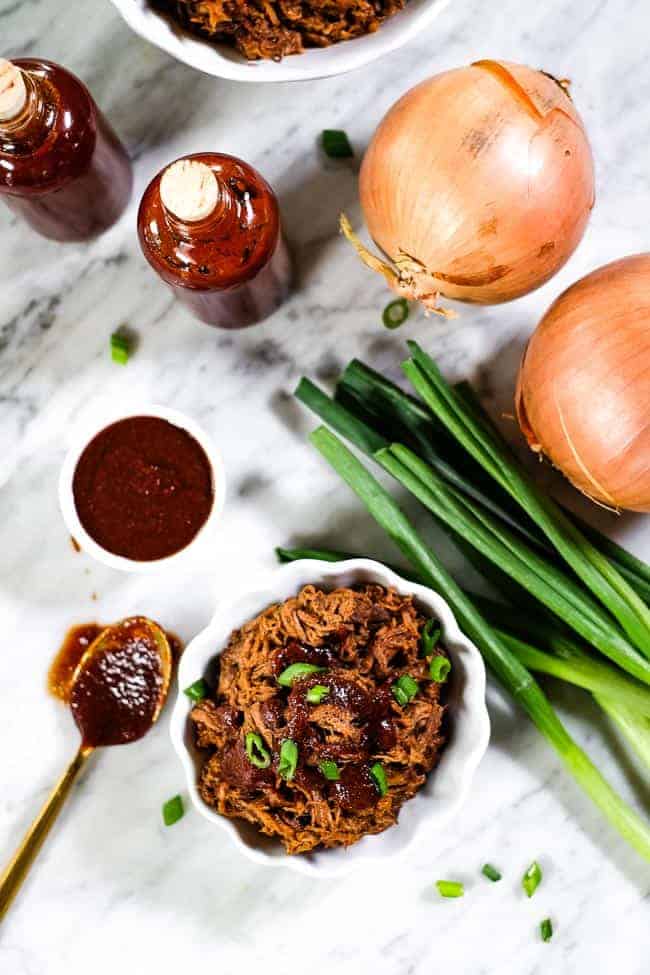 Slow cooker BBQ beef in a ramekin with BBQ sauce and chopped green onion on top. Extra sauce and onions on the table.