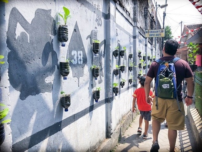 A man and a young boy walk through a small walkway in Yogyakarta, Indonesia, where travelers can learn Indonesian. To the left of them, they pass a wall decorated with plastic bottles that have been converted into planters, and paintings of shadow puppets on the wall.