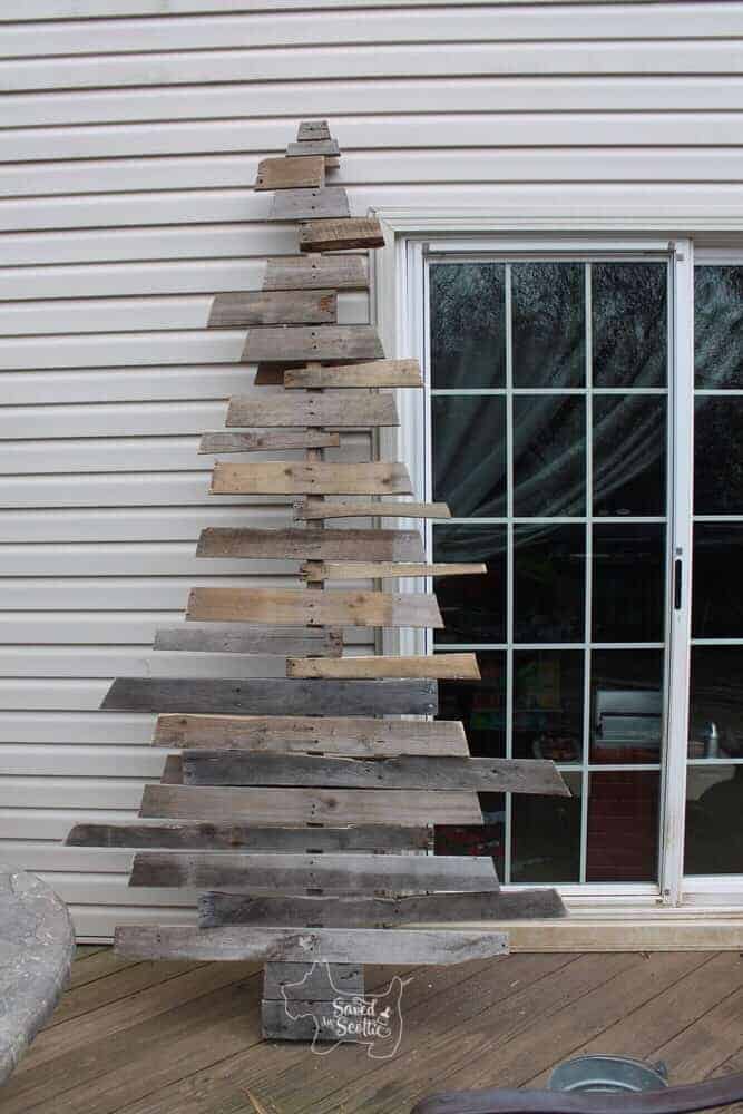 a finished version of a wood pallet christmas tree that is full and randomly uneven on the sides leaning against the siding on a house next to a glass sliding door.