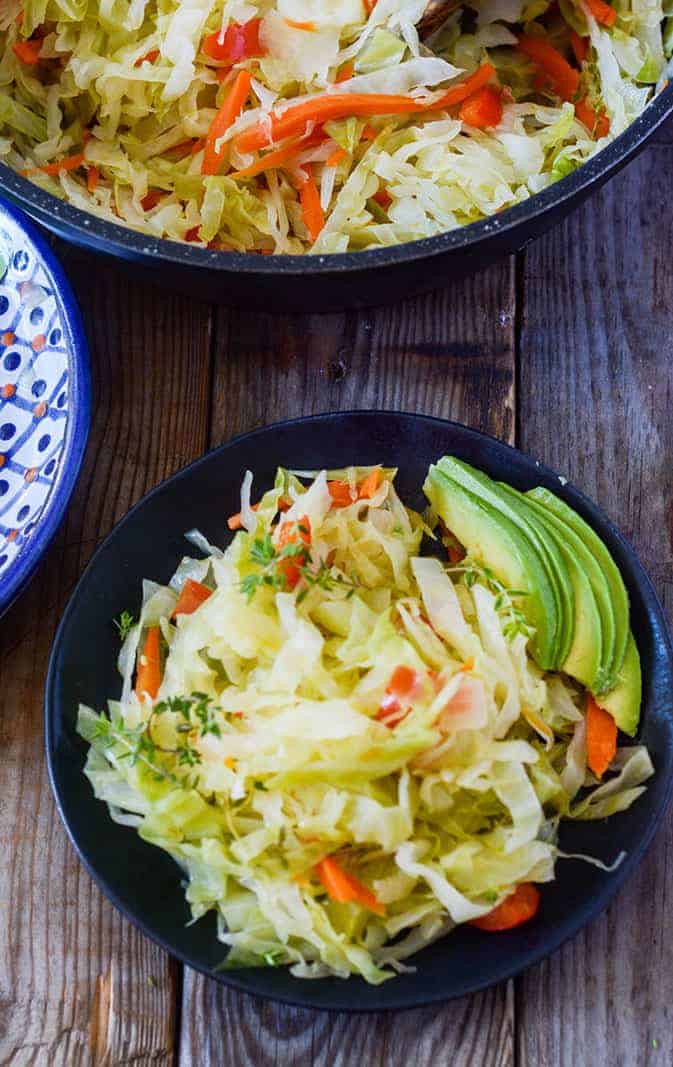 Jamaican Steamed Cabbage on a black plate, garnished with avocado on a wooden background