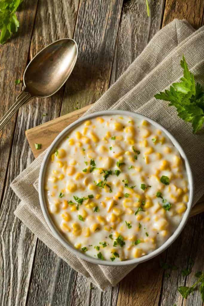 2 Creamed Corn Recipes (with & without cream) A bowl of creamed corn on a brown napkin and a wooden board
