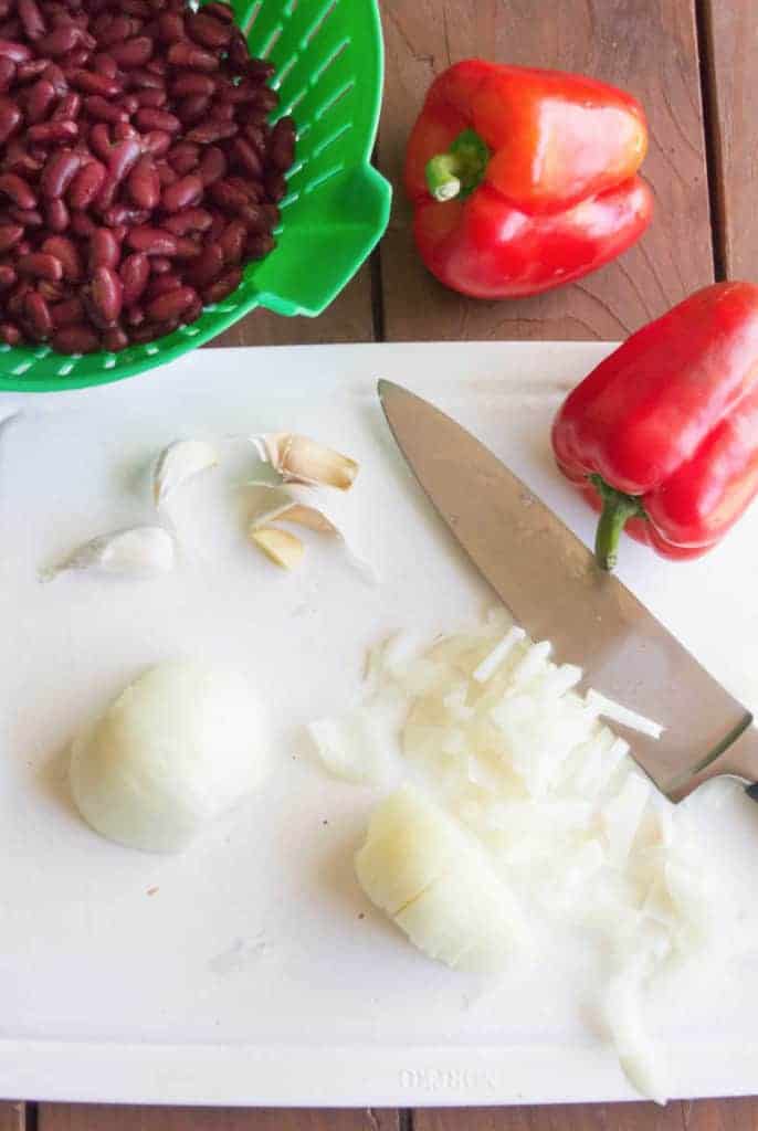 An onion is chopped on a white cutting board. A knife rests to the right. Garlic, two red peppers and beans in a green strainer sit nearby.