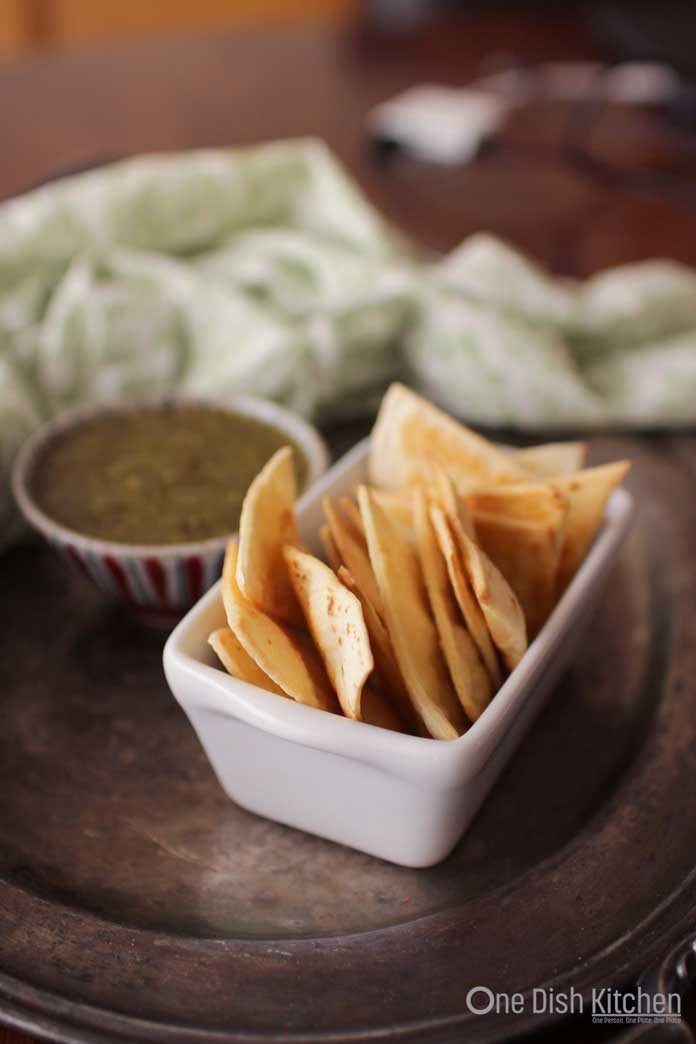 A small bowl of homemade tortilla chips next to a small bowl of green salsa all on a metal tray