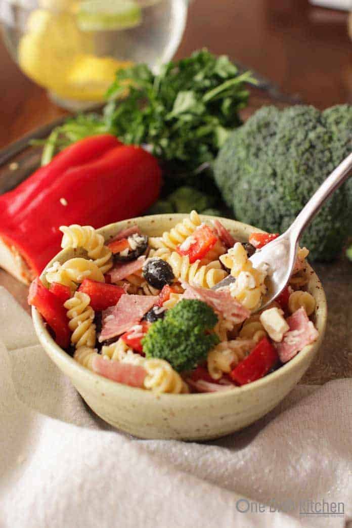 A forkful of italian pasta salad from a small bowl on a metal tray with a red pepper and a head of broccoli