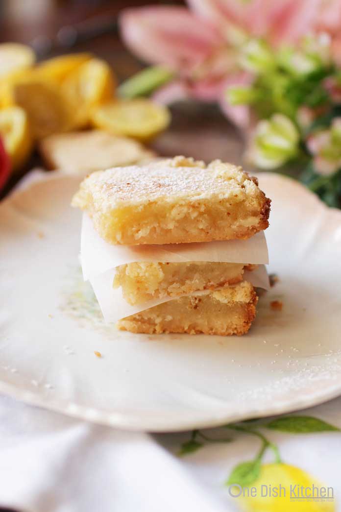 A stack of three lemon bars on a plate with lemon slices and pink flowers in the background