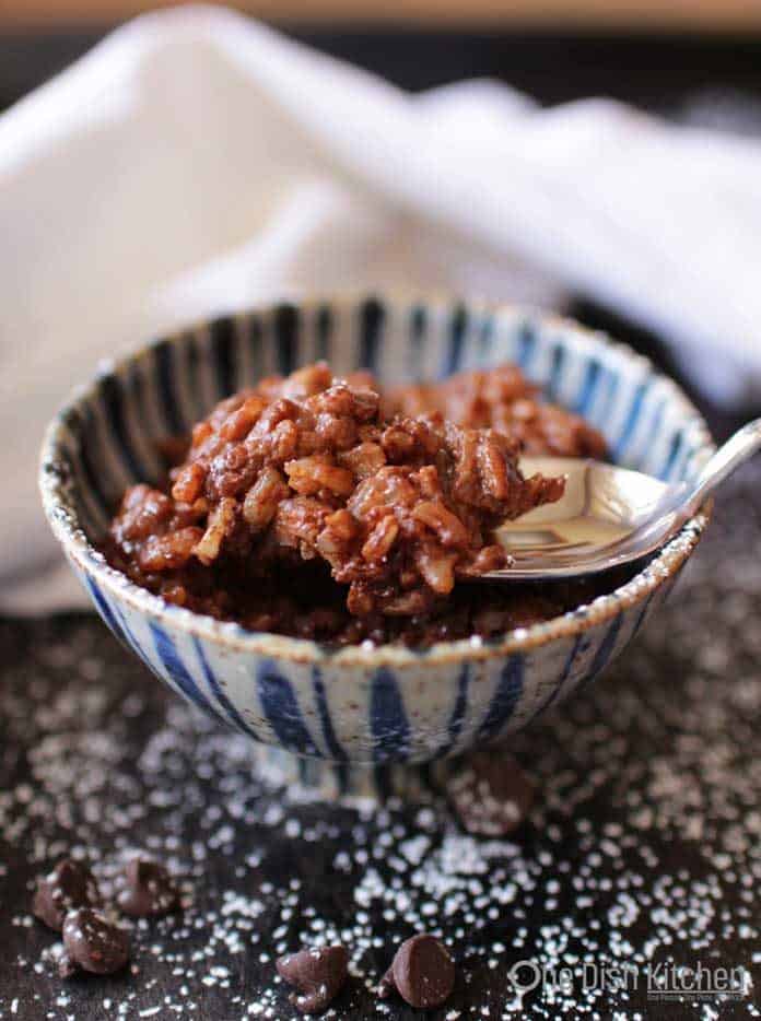A spoonful of Chocolate rice pudding from a bowl on a dark surface with scattered chocolate chips and powdered sugar