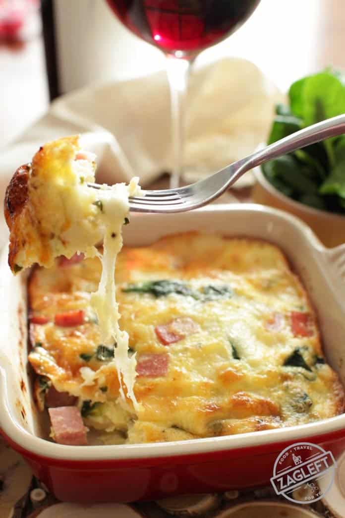 A forkful of crustless quiche with cheese, spinach, and ham from a small baking dish next to a glass of red wine