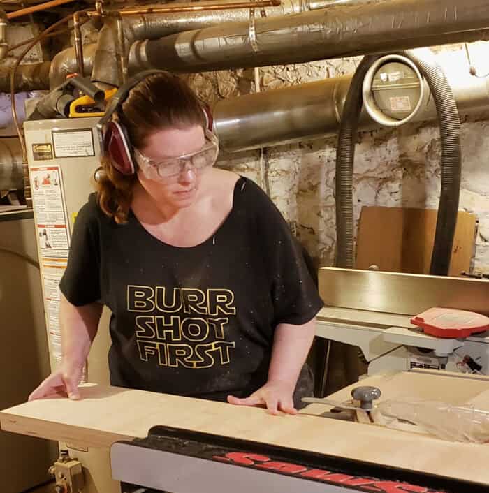 Amy, wearing ear protection, moving a wooden board though a table saw