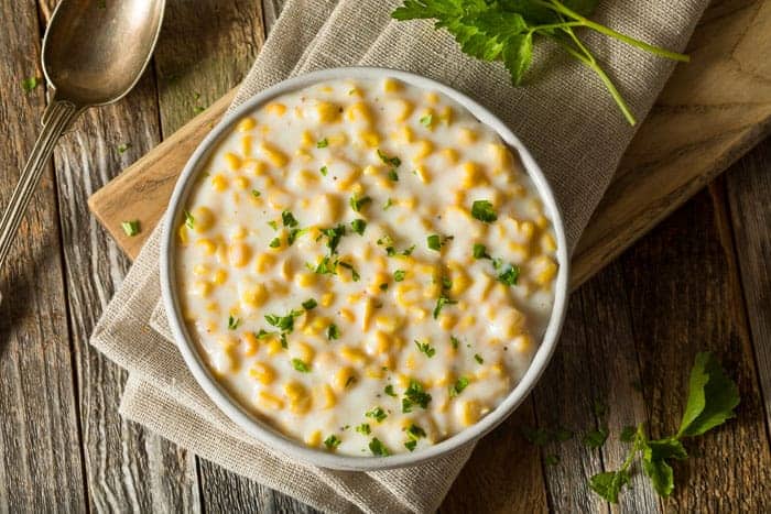 2 Creamed Corn Recipes (with & without cream) A bowl of creamed corn on a brown napkin and a wooden board
