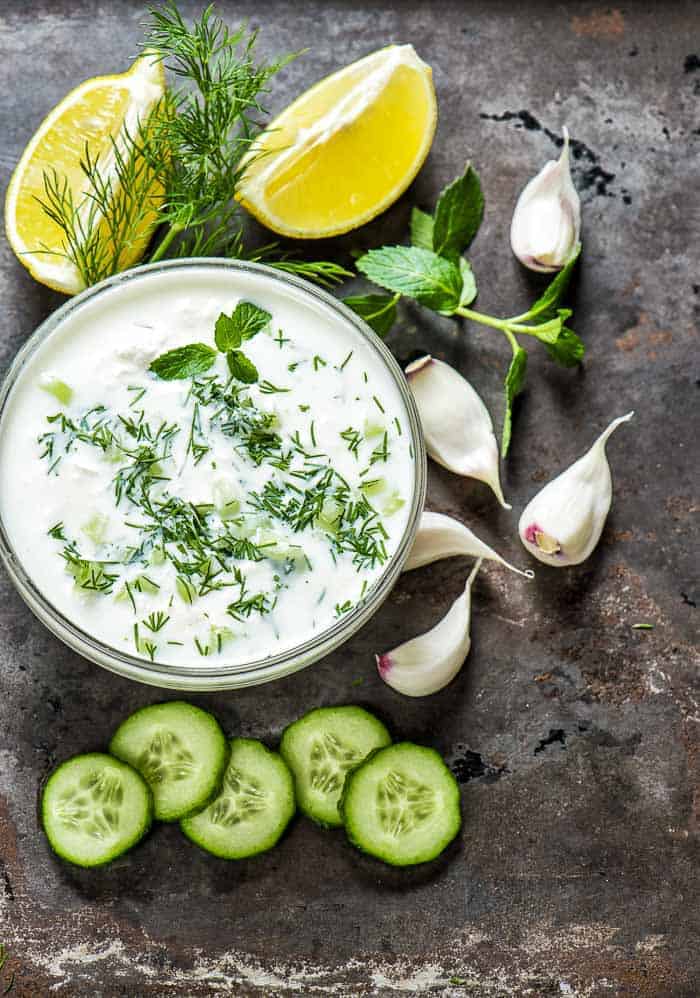Tzatziki Sauce Recipe a bowl of tzatziki sauce with fresh lemon, fresh cucumbers, garlic cloves, and fresh dill and mint around the bowl on a table