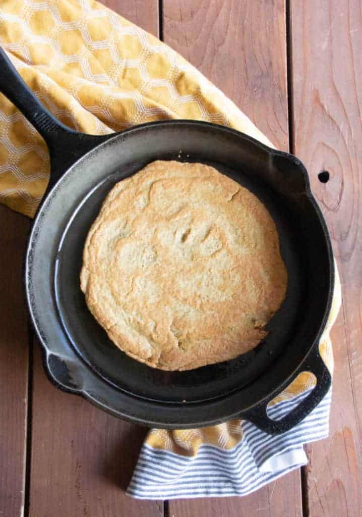 A lentil and chickpea flatbread sits inside a black cast iron pan, resting on a yellow tea towel atop a wooden table surface.