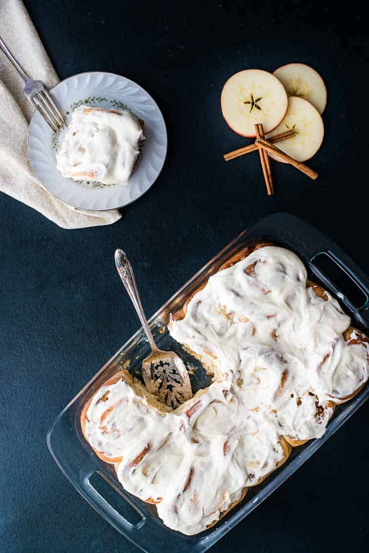 A pan of apple cinnamon rolls with vanilla cream cheese frosting next to a single roll on a plate.