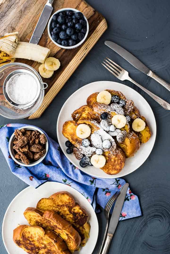 A serving of vanilla french toast next to a serving plate and bowls of candied pecans, blueberries, powdered sugar, and a banana.