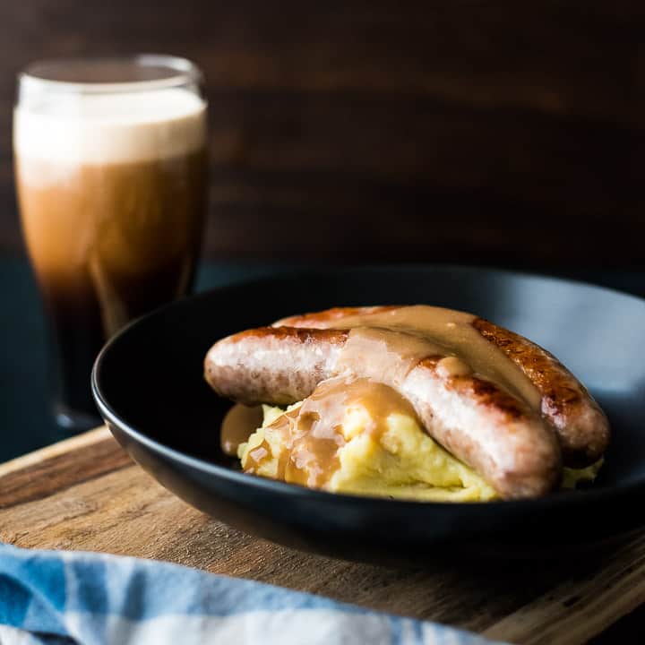 Bangers and mash with Guinness gravy in a dark bowl with a freshly poured Guinness in the background.