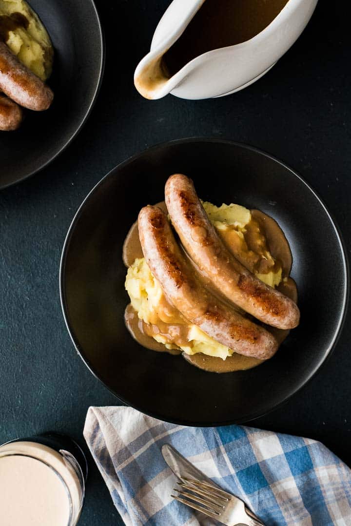 Overhead shot of bangers and mash with Guinness gravy plated in two bowls with a gravy bowl and a freshly poured Guinness.