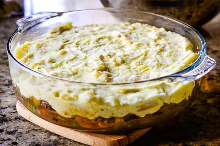 A round, glass baking dish with the meat, vegetables, and gravy combination layered in the bottom and mashed potatoes on top, ready to be baked.
