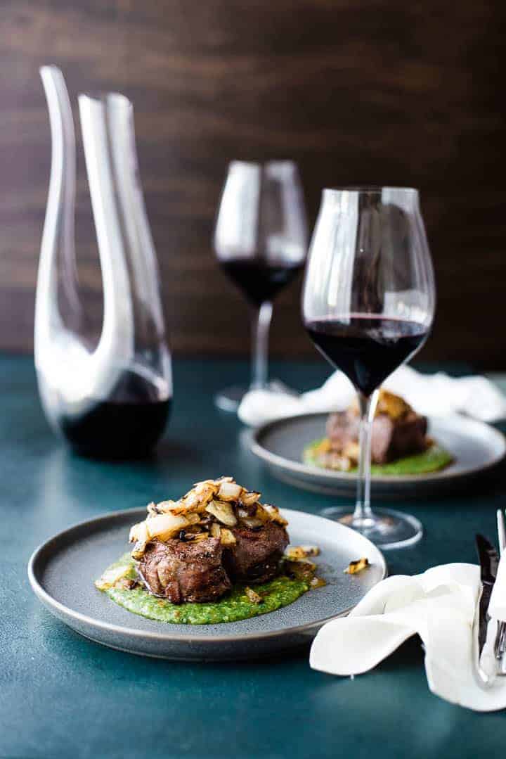A thick cut filet mignon, resting on a green colored peach mango cilantro sauce and topped with grilled onions. A second serving is in the background along with a decanter of red wine and two glasses.