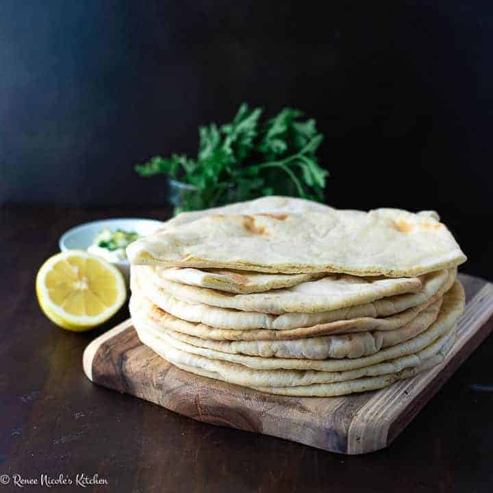 A stack of thin puffed pita bread on a wooden cutting board next to a bowl of tzatziki sauce, a sliced lemon, and a bunch of parsley.