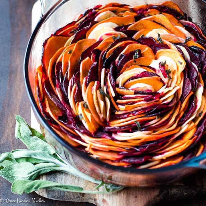 Close up square shot of oven roasted root vegetables (beets, sweet potatoes, & parsnips) in a glass pan next to a sprig of sage.