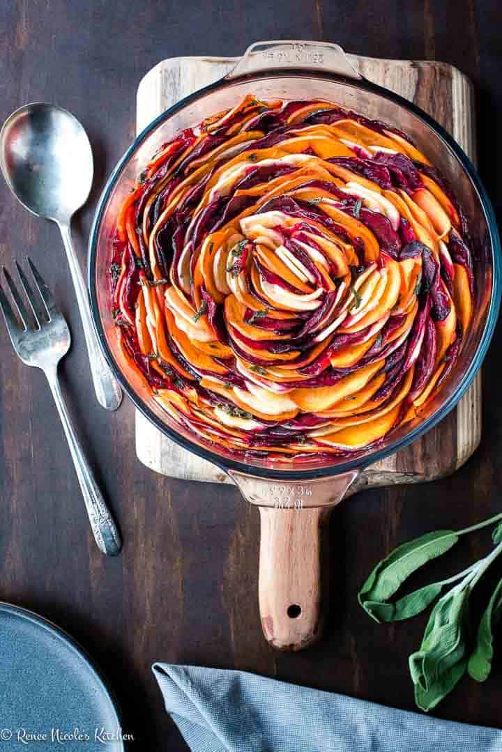 Overhead shot of oven roasted root vegetables in a glass pan on a wooden cutting board. Antique silver serving spoons to the left, a bowl and napkin to the bottom, and a sprig of sage.