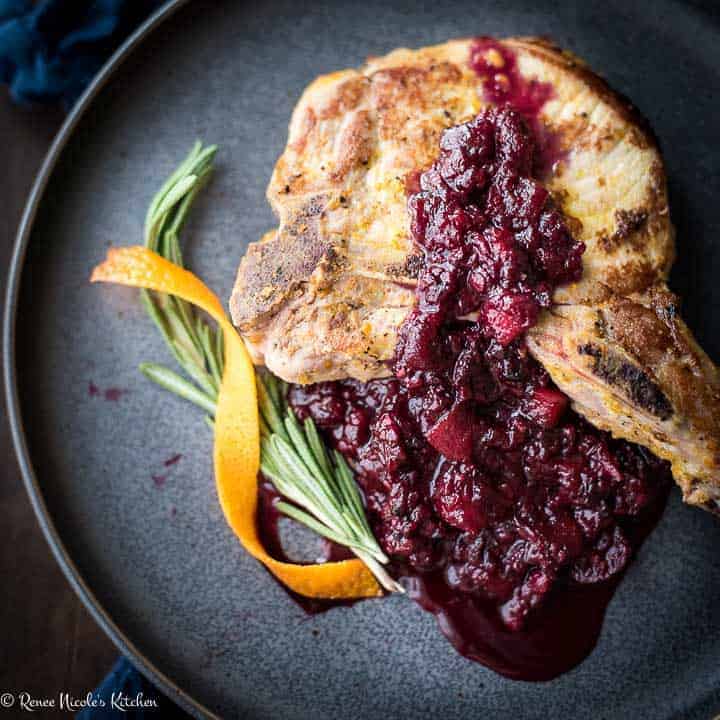 Close up, square image of a bone in pork chop on a gray plate with a spring of roemary and strip of orange zest to the side, topped with blackberry applesauce.