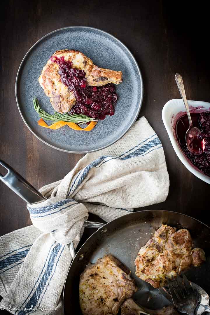 Overhead shot of a serving of pork chops and applesauce on a gray plate next to a white container of blackberry applesauce and a skillet of pan seared oven roasted pork chops.