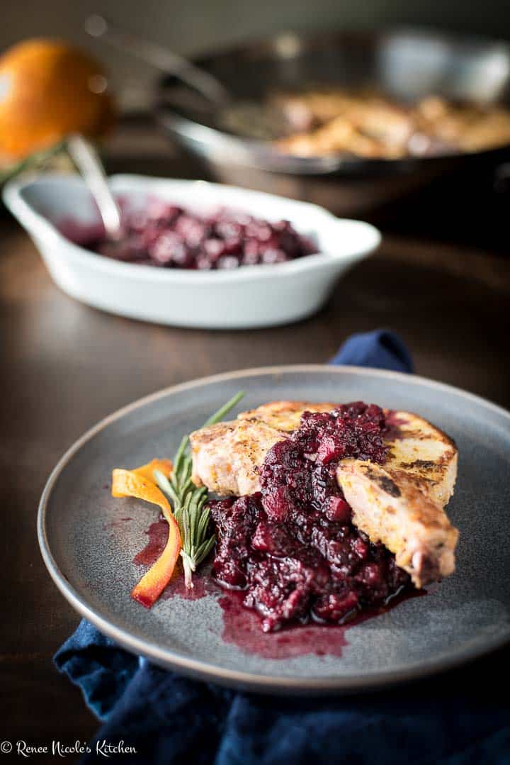 A serving of pork chops and blackberry applesauce on a gray plate in front of a serving bowl of blackberry applesauce and a skillet of pan seared pork chops.