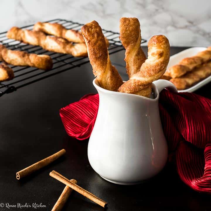 Square image of cinnamon twists in a white jug, with cinnamon sticks scattered in the front and more cinnamon sticks on a cooling rack in the background.
