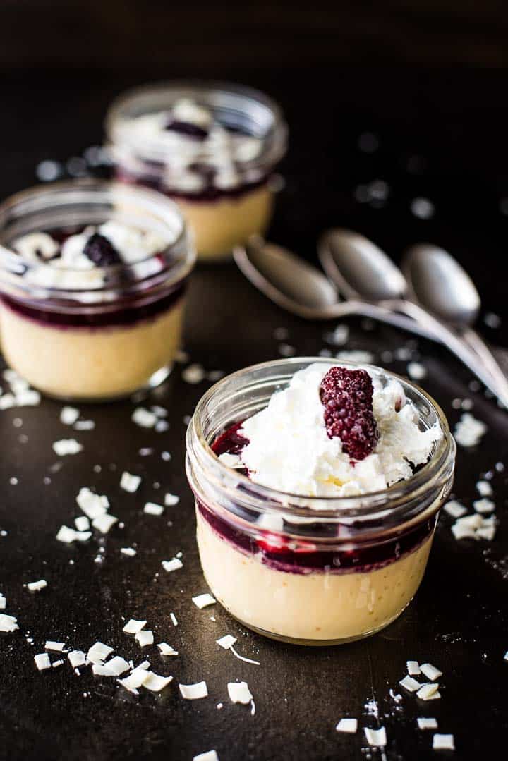 Blackberry white chocolate pots de creme in a glass jar with whipped cream and a blackberry on top with two additional servings in the background.