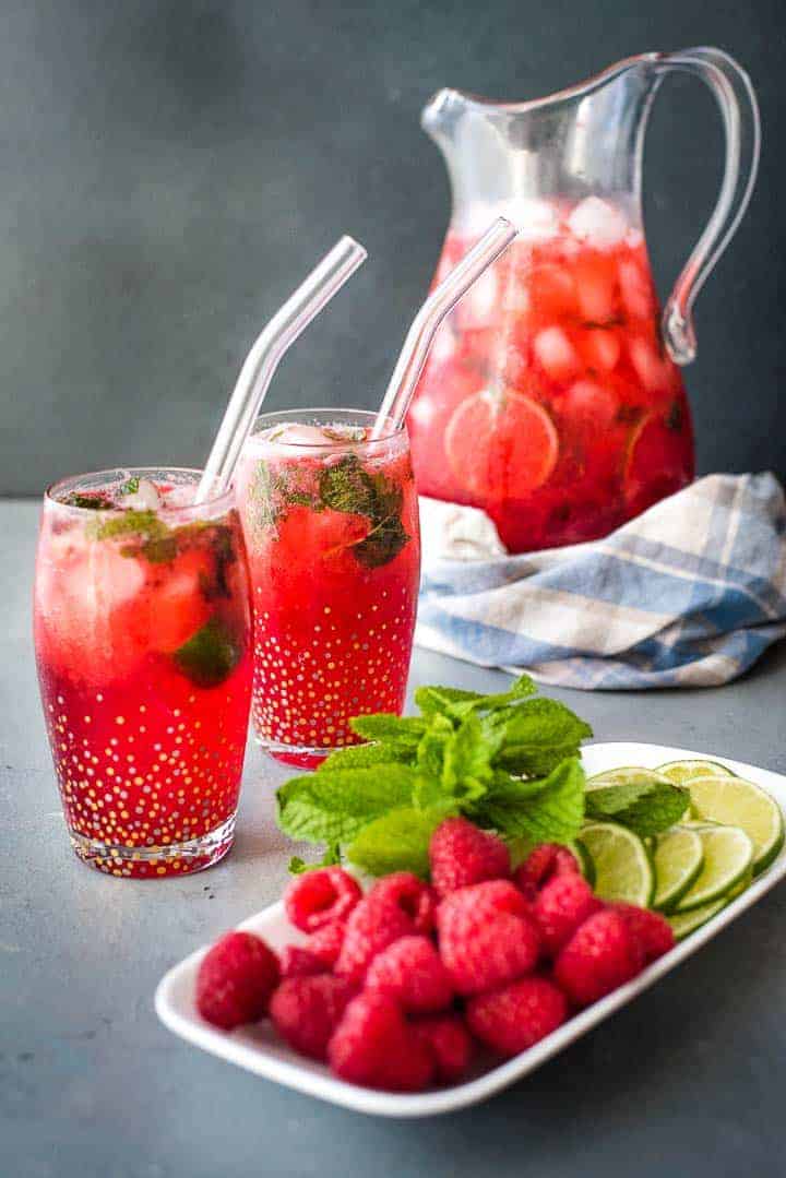 Two raspberry mojito cocktails with a garnish tray in the front and a pitcher of mojitos in the back.