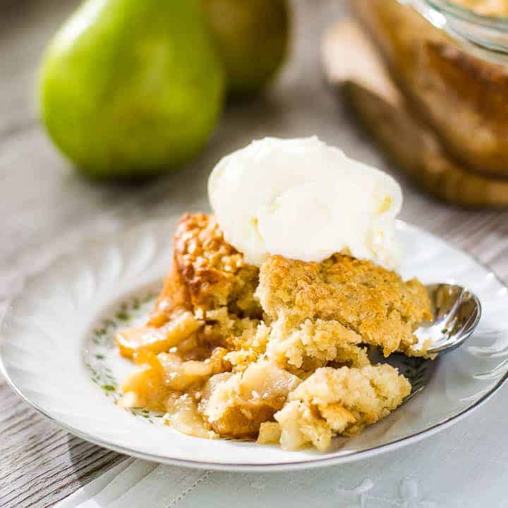 A close-up of a single serving of spiced pear cobbler with a dollop of sweet cream ice cream on top and two pears in the background.