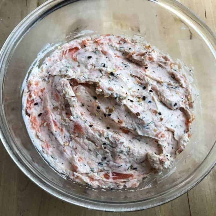 clear bowl containing everything bagel and lox spread on wood cutting board