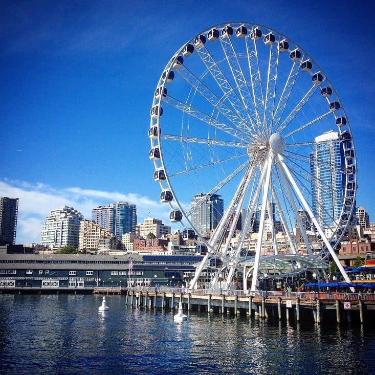 A large white ferris wheel sits on a dock, with a city skyline in the background. This is the Seattle Great Wheel, one of the activities in Seattle for families and tourists.