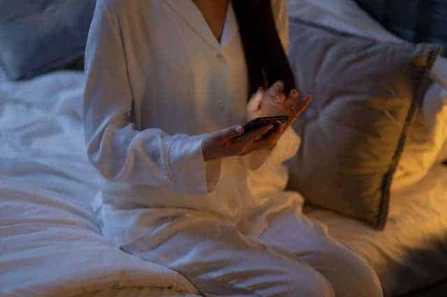 Une femme en pyjama, avec un smartphone à la main, est assise sur le lit