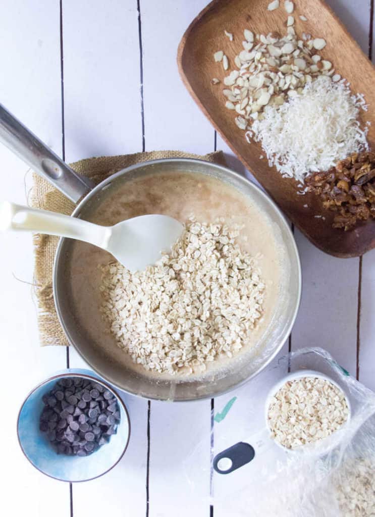 Almond butter, coconut oil and honey have been combined together in a small pot and warmed until everything is melted. Oats have been added to the top of the mixture. A small blue ingredient bowl with choloate chips, and wooden tray of ingredients sits nearby.