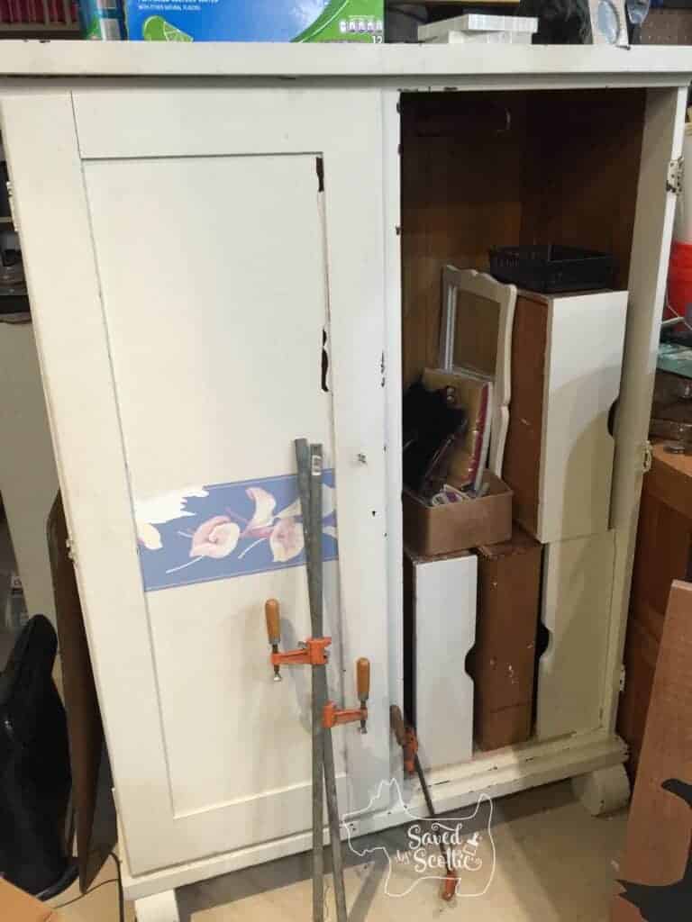 an old armoire with a broken door on the right side. Covered in chipping paint and wallpaper border. The open side is full of the drawers and a mirror. Two long clamps are leaning against the front of the armoire