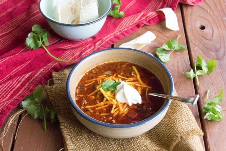 A white, blue rimmed bowl sits on a burlap napkin. The bowl is full of perfect chili, topped with shredded cheese, sour cream and garnished with cilantro.