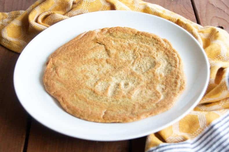 On top of a brown wood surface, a cooked lentil and chickpea flatbread sits on a white plate, a yellow tea towel is nearby.