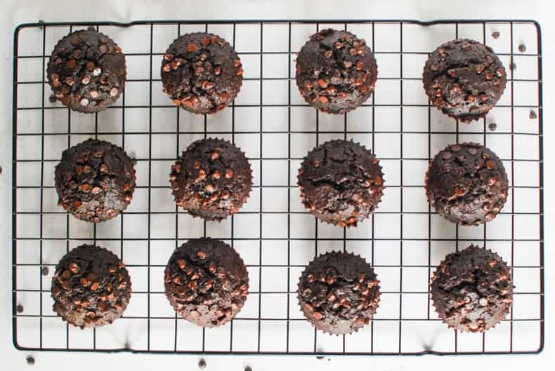 Overhead shot of a dozen coconut flour chocolate muffins on a black cooling rack and white surface,
