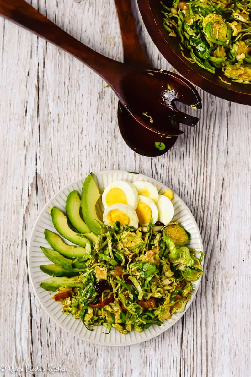Overhead shot showing a serving of shaved brussels sprouts salad next to the serving bowl of shaved brussels sprouts salad with salad servers in the middle.