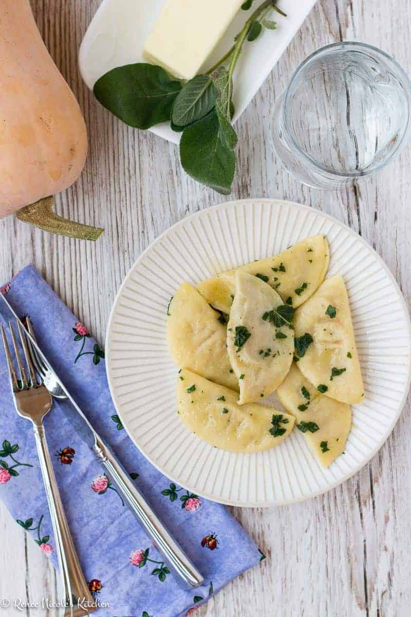 An overhead shot of plated butternut squash ravioli served on a white plate with sage brown butter sauce. Also showing a butternut squash, a block of butter and a sprig of sage.