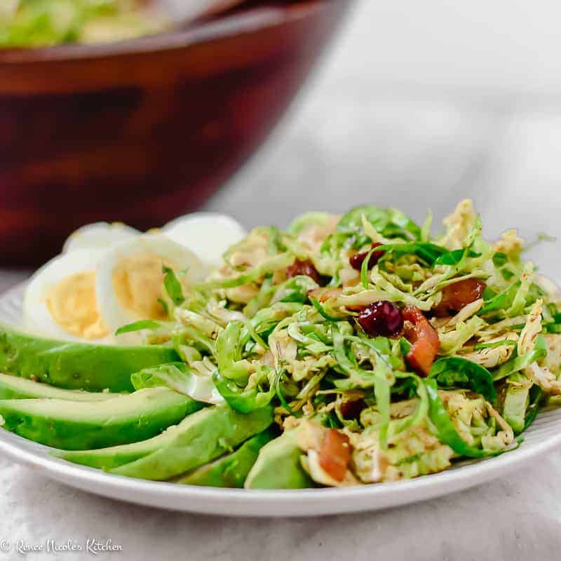A close up of shaved brussels sprouts salad on a lined white plate with sliced avocado and sliced hard boiled egg.