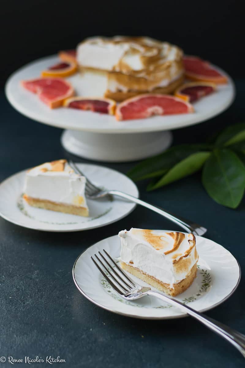 Two slices of citrus dream tart on white bowls in front of the rest of the tart in the background on a white marble cake stand.
